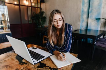 Lovable girl writing something in her notebook. Indoor shot of adorable long-haired woman using white laptop in cafe.